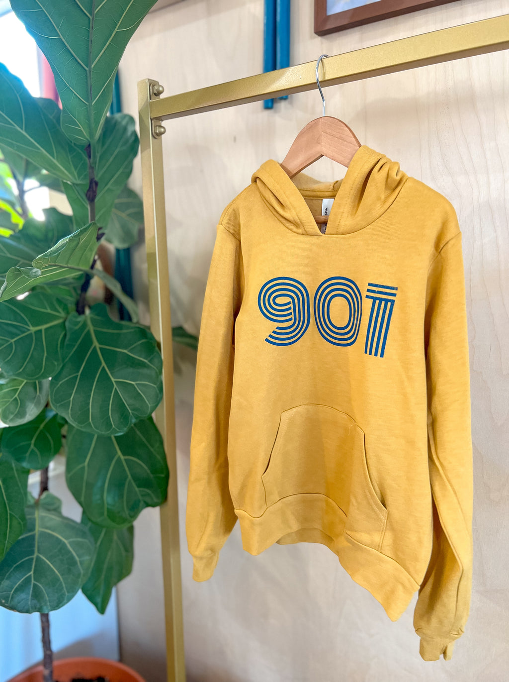 A mustard yellow hoodie with the text '901' and a blue stripe design hanging on a hanger against a backdrop of green foliage.