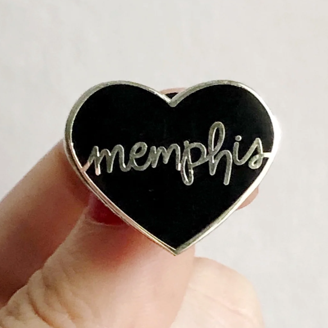 hand holding black enamel pin with silver memphis lettering on white background