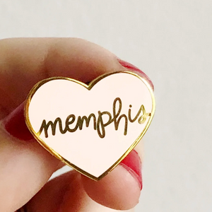 hand holding pink heart enamel pin with memphis written in gold cursive on cream background